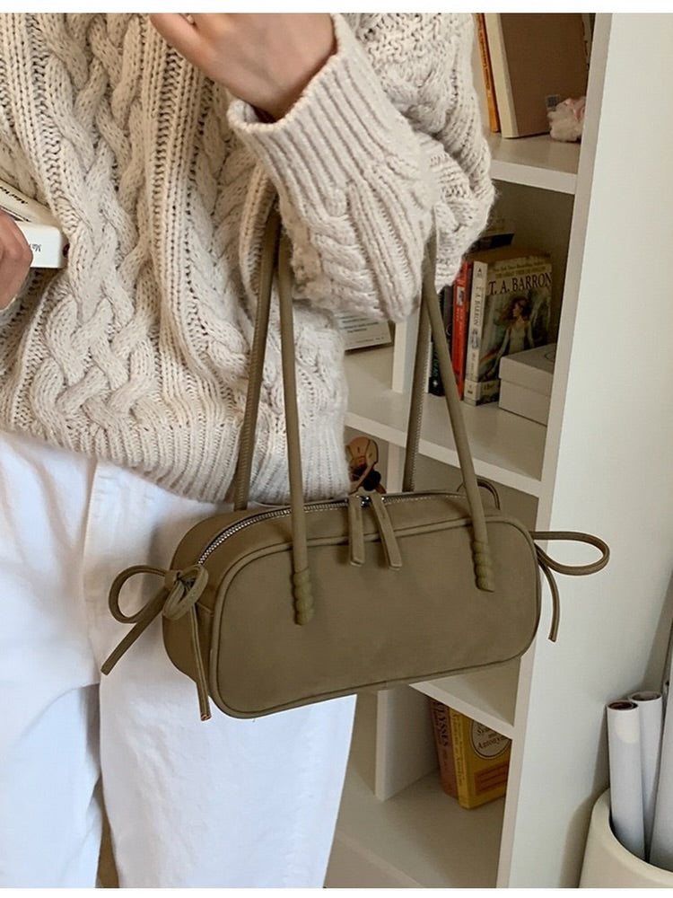 A person wearing a cream cable-knit sweater and white pants carries a taupe-brown faux-leather shoulder bag with a top zipper, tassel detail, and bow straps, set against a white bookshelf background.
